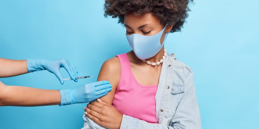 Curly haired young African American woman gets vaccine in shoulder prevents from coronavirus wears protective mask poses against blue background. Immunization campaign and health care concept