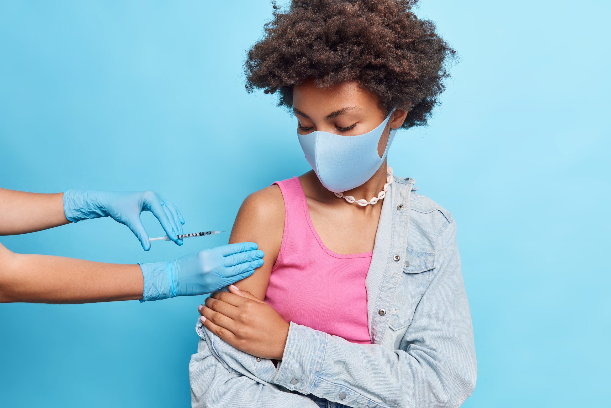 Curly haired young African American woman gets vaccine in shoulder prevents from coronavirus wears protective mask poses against blue background. Immunization campaign and health care concept